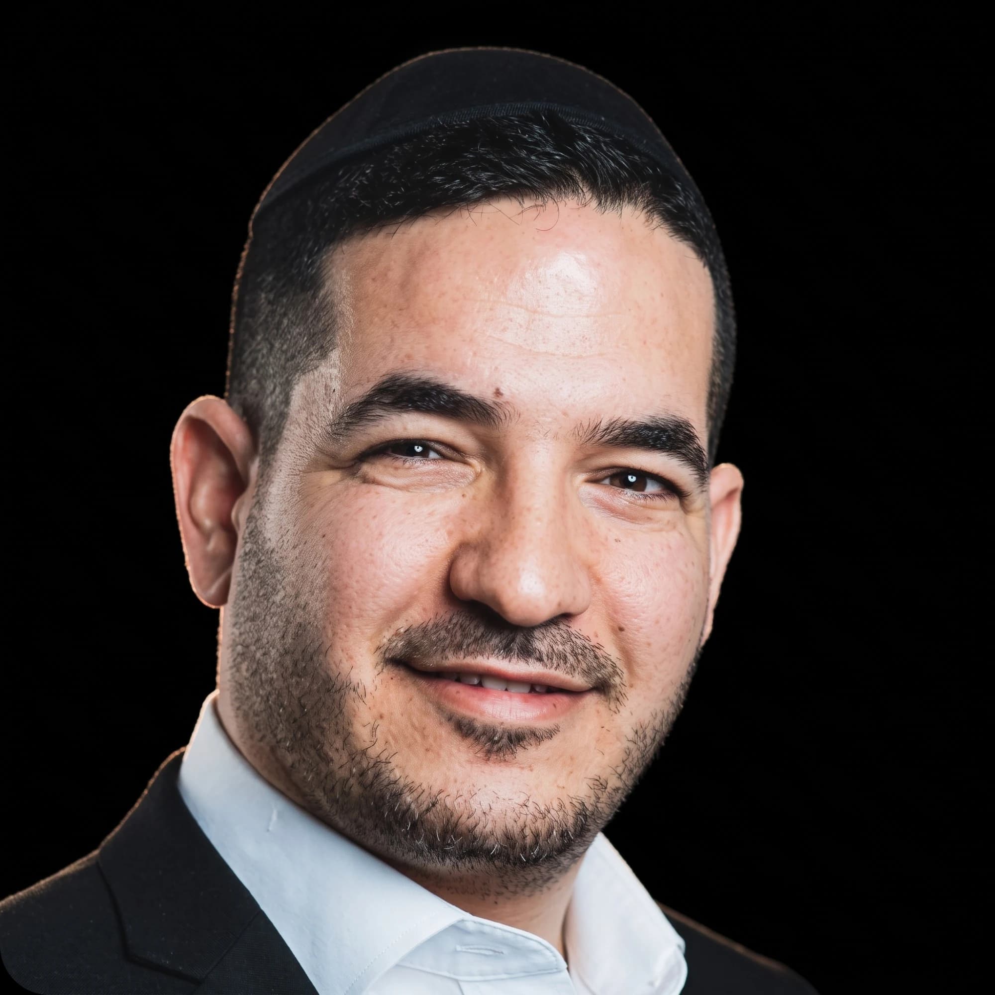 פורטרט של יוסף בן שושן מחייך, בחליפה כהה, חולצה לבנה וכיפה שחורה